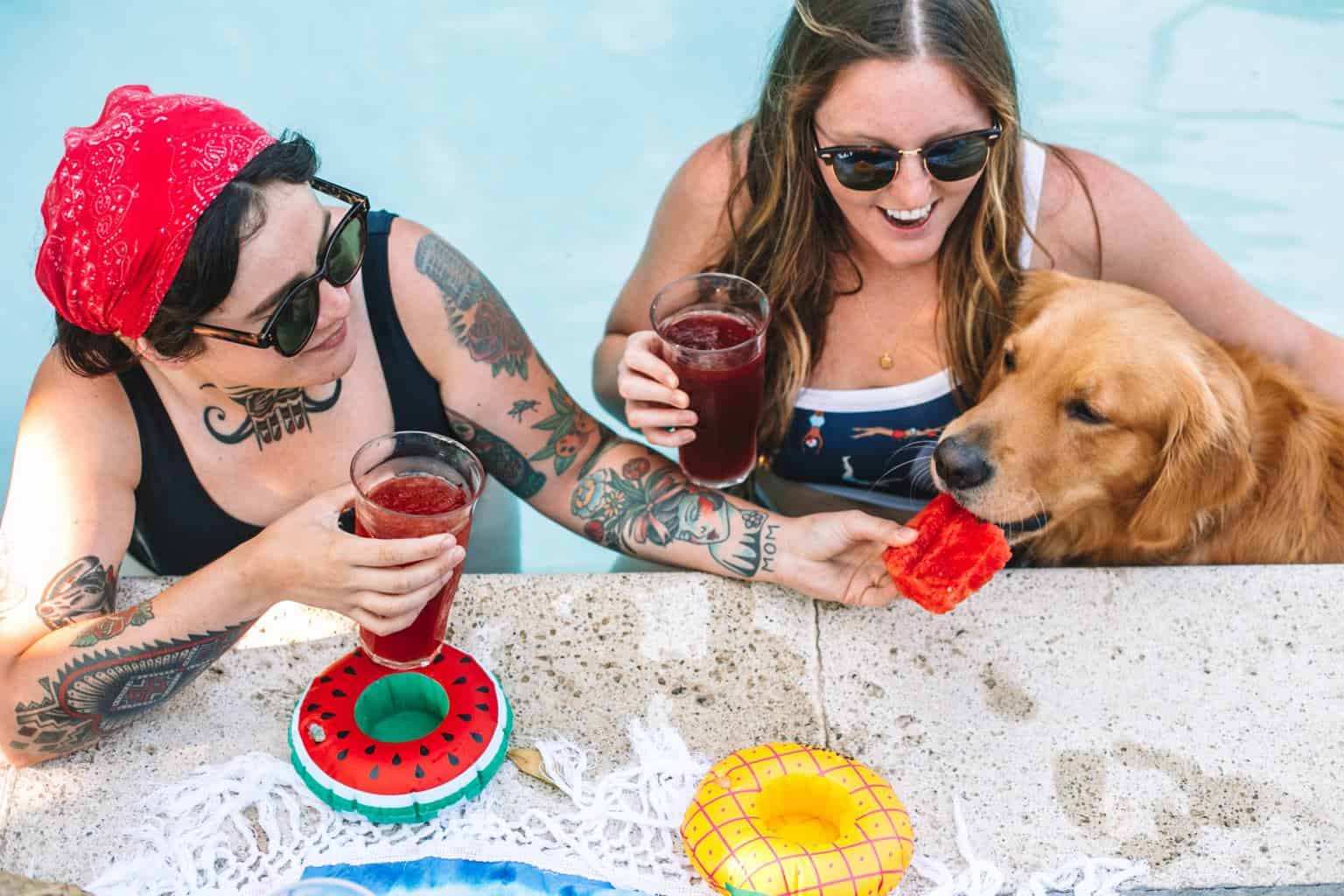 Two girls inside of a swimming pool feeding a dog a piece of watermelon. 