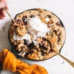 hand scooping skillet chocolate chip cookie topped with vanilla ice cream wrapped in gold kitchen towel