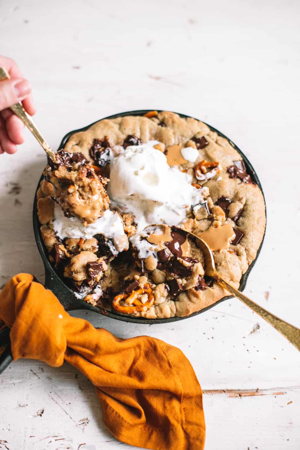 hand scooping skillet chocolate chip cookie topped with vanilla ice cream wrapped in gold kitchen towel