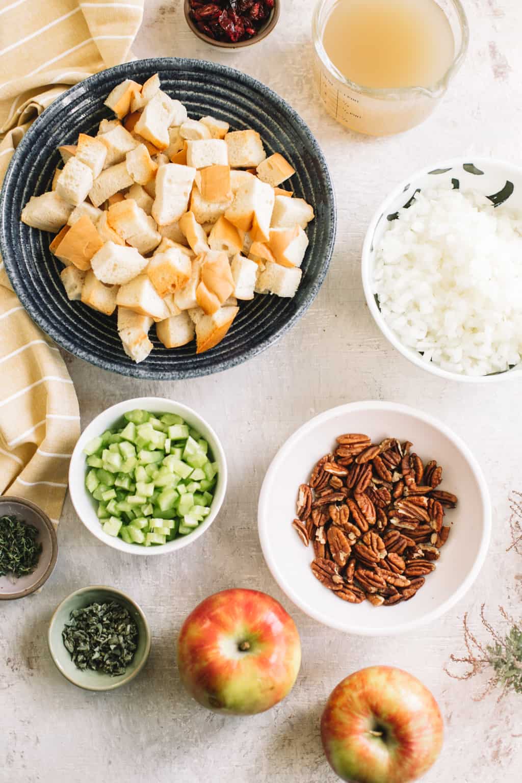 bread cubes in black bowl, chopped onions and celery in two white bowls, pecans in white bowl with apple and chopped herbs