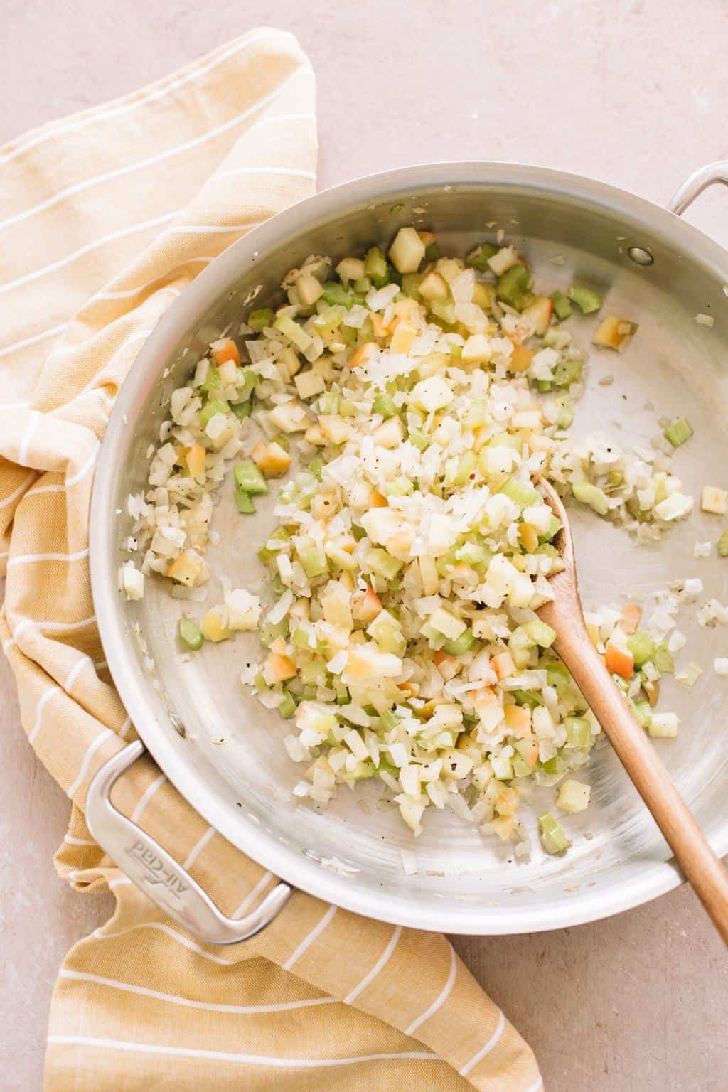 onions, celery and apples sauteing in large pan with wooden spoon on brown kitchen towel