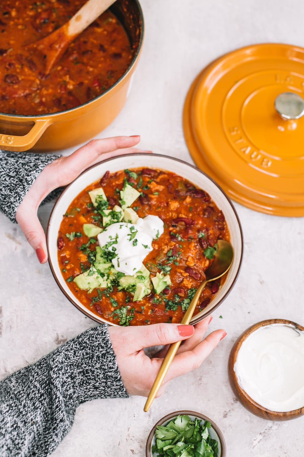 two hands holding chipotle turkey chili recipe in white bowl with gold spoon with remanining turkey chili in yellow dutch oven