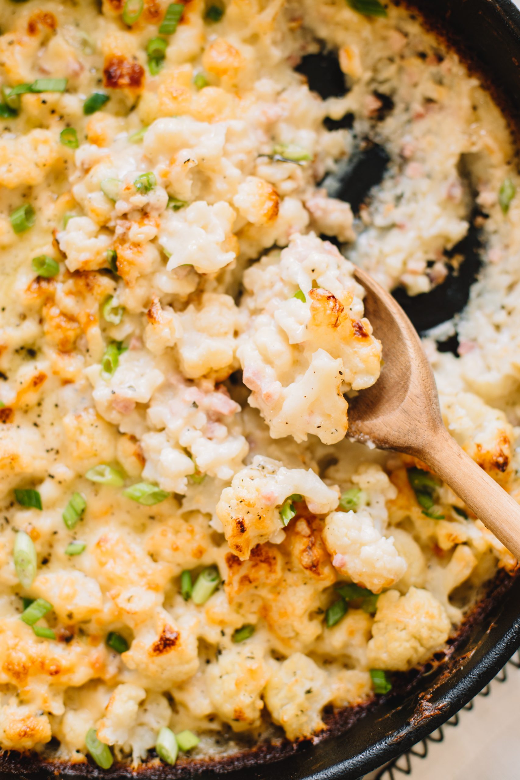 wooden spoon scooping easy cheesy cauliflower bake with pancetta in cast iron skillet