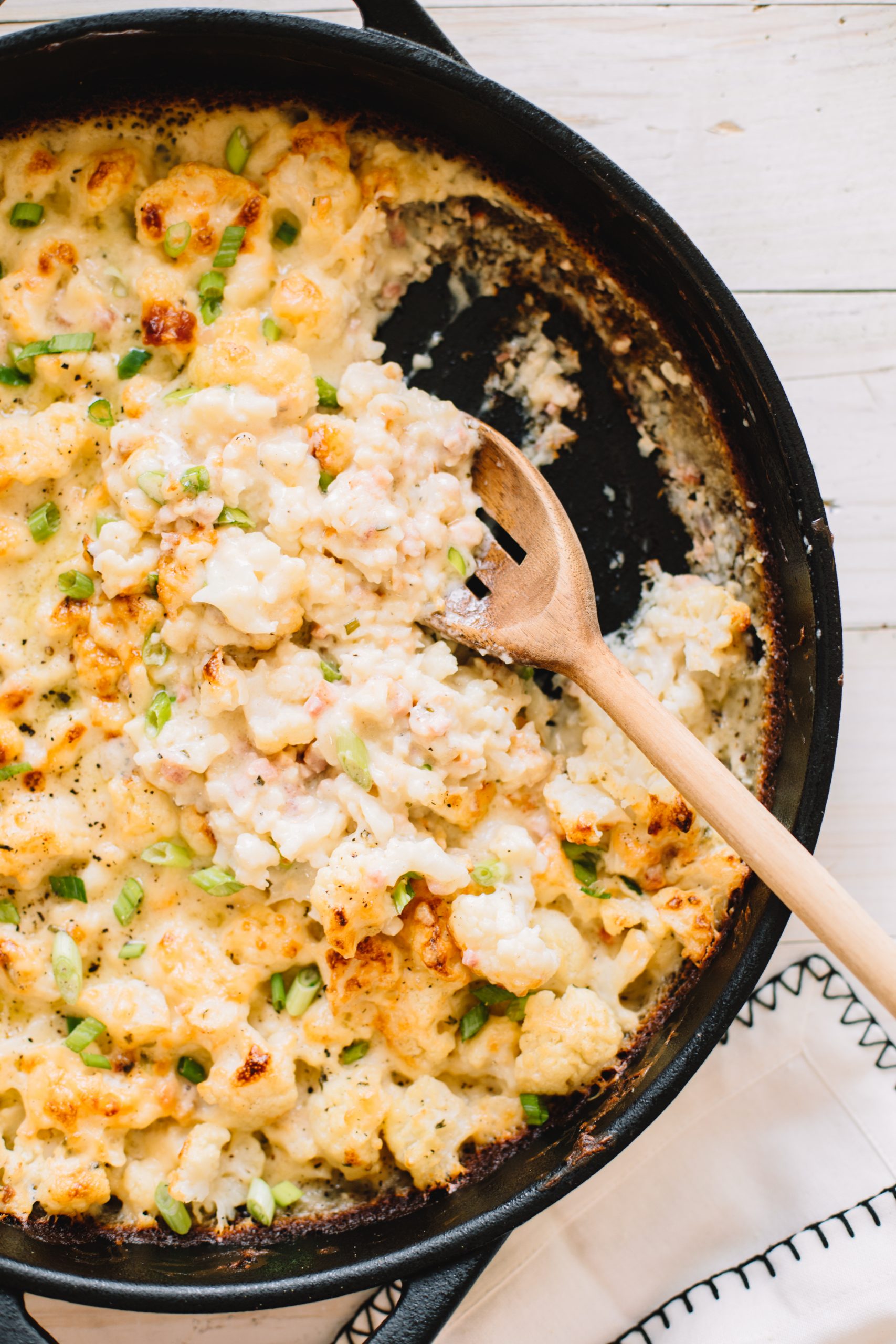 cheesy cauliflower casserole in cast iron skillet being scooped with wooden spoon