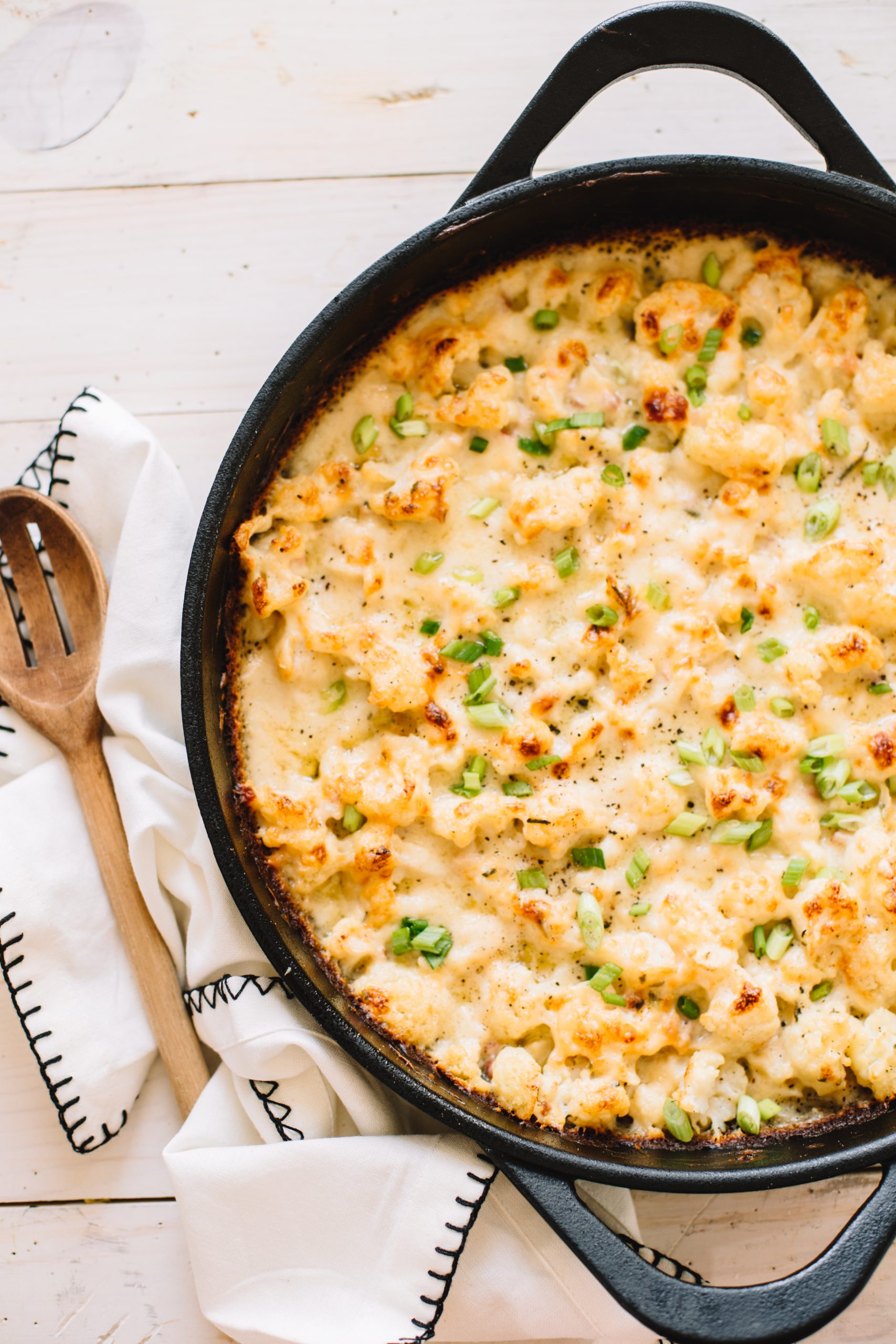 cheesy cauliflower bake with pancetta in cast iron skillet with wooden spoon on kitchen towel