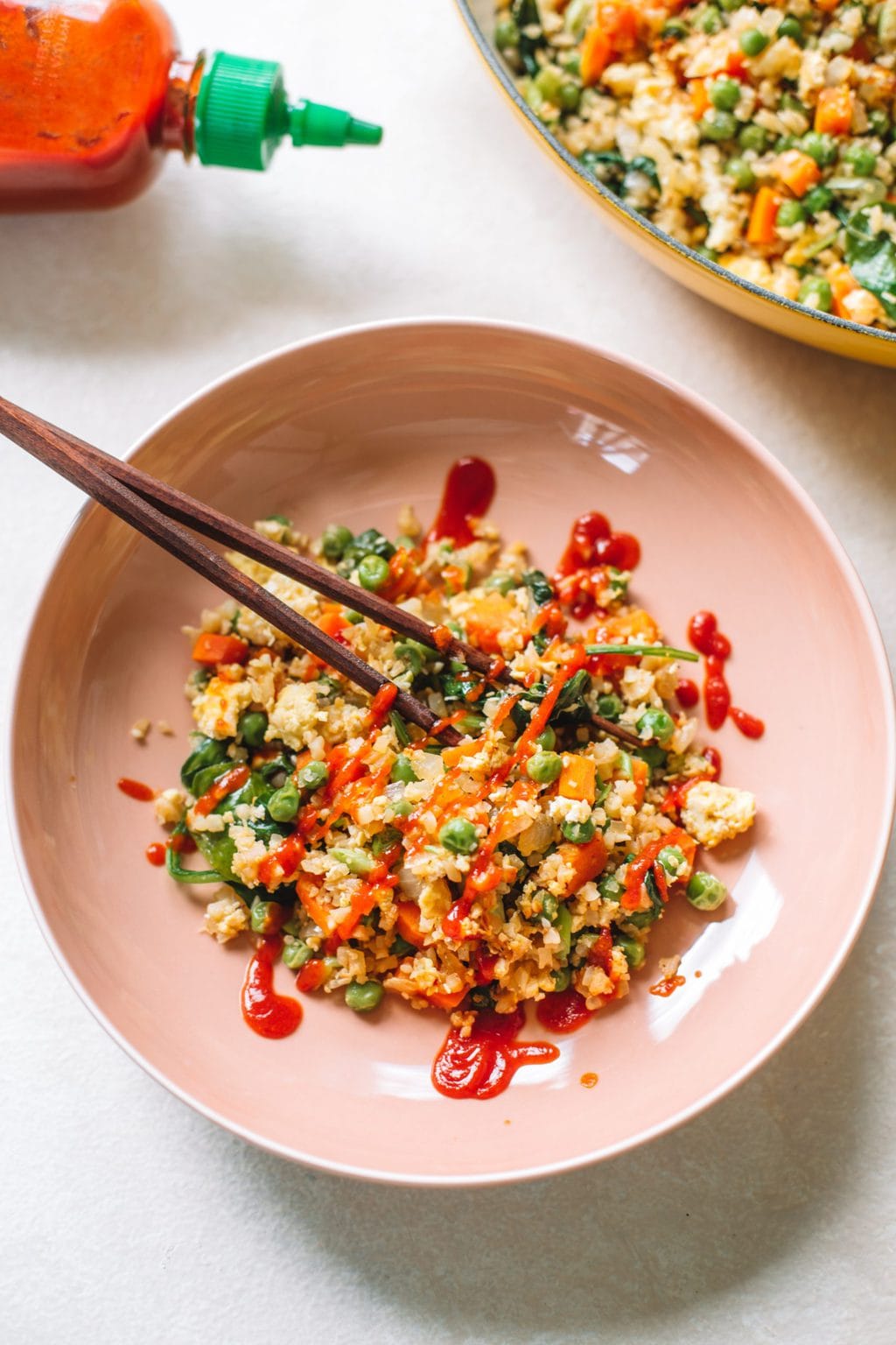 A pink bowl filled with cauliflower fried rice, topped with sriracha and served with chop sticks. 