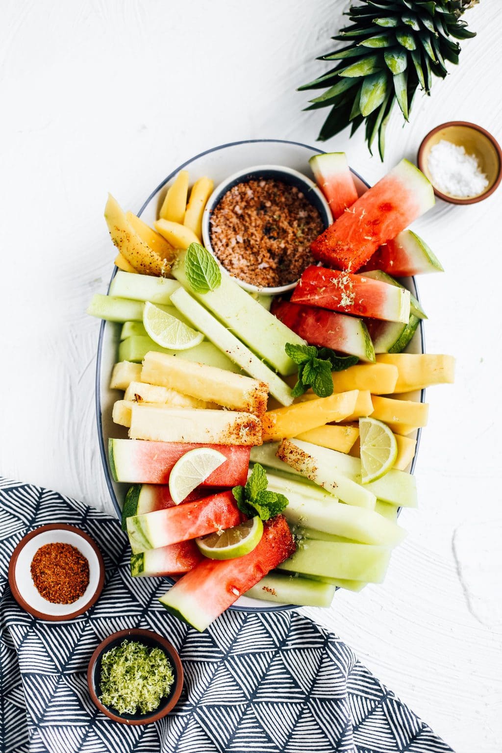 Honeydew, watermelon, pineapple and mango spears on an oblong platter with a bowl of chili lime seasoning.