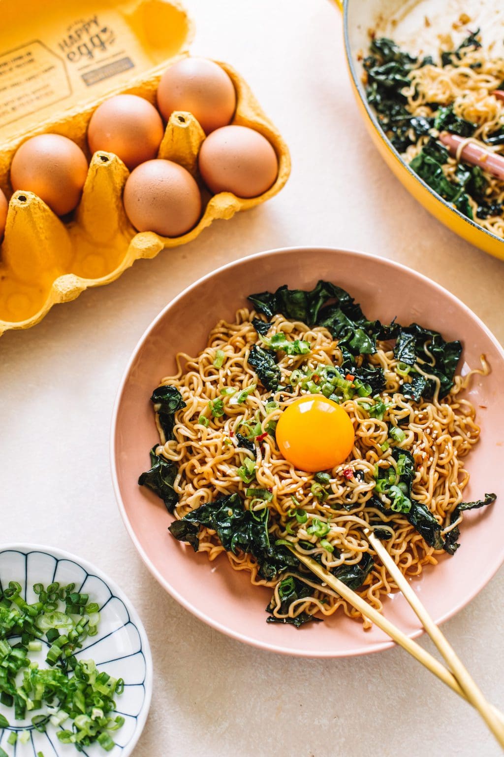 A pink bowl of garlic butter ramen noodles with a carton of yellow eggs. 