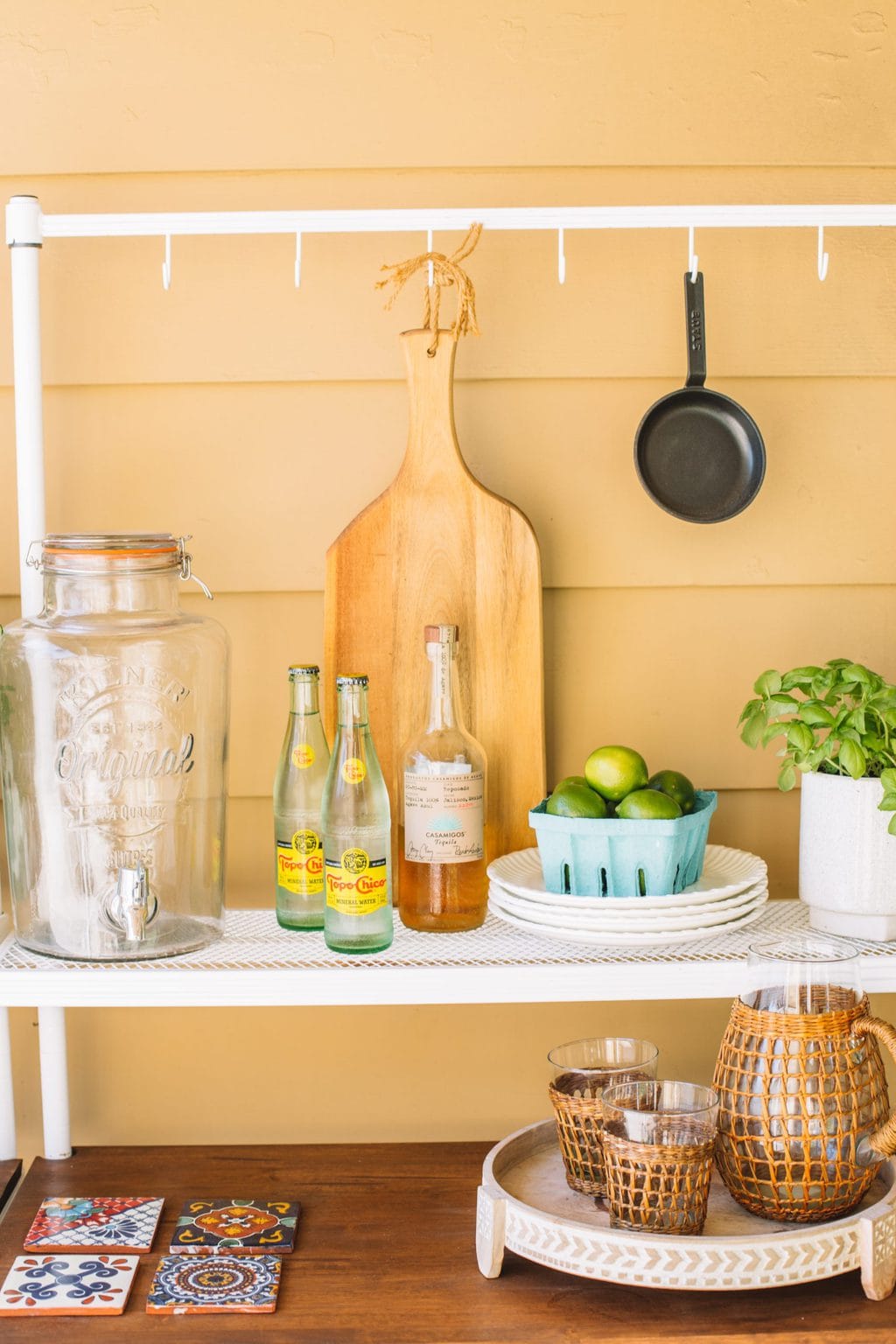 An outdoor patio buffet setup with a drink dispenser, sparkling water, tequila, limes and outdoor plates.