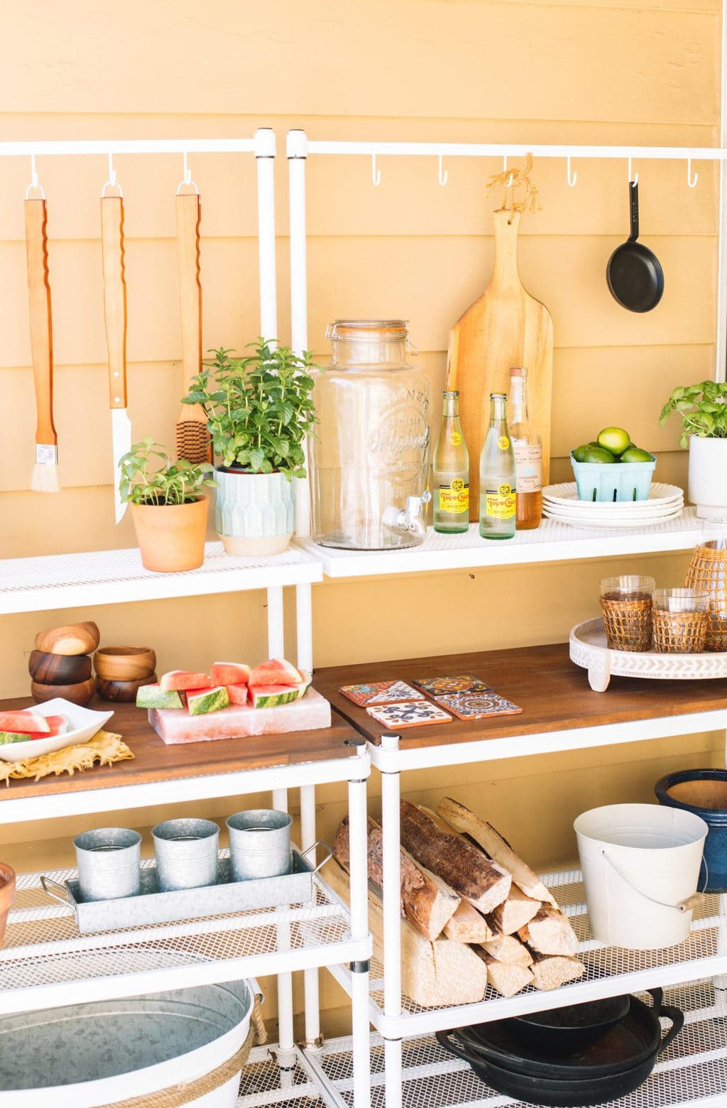 An outdoor patio buffet setup with bbq tools, plates, cups, planters and firewood.