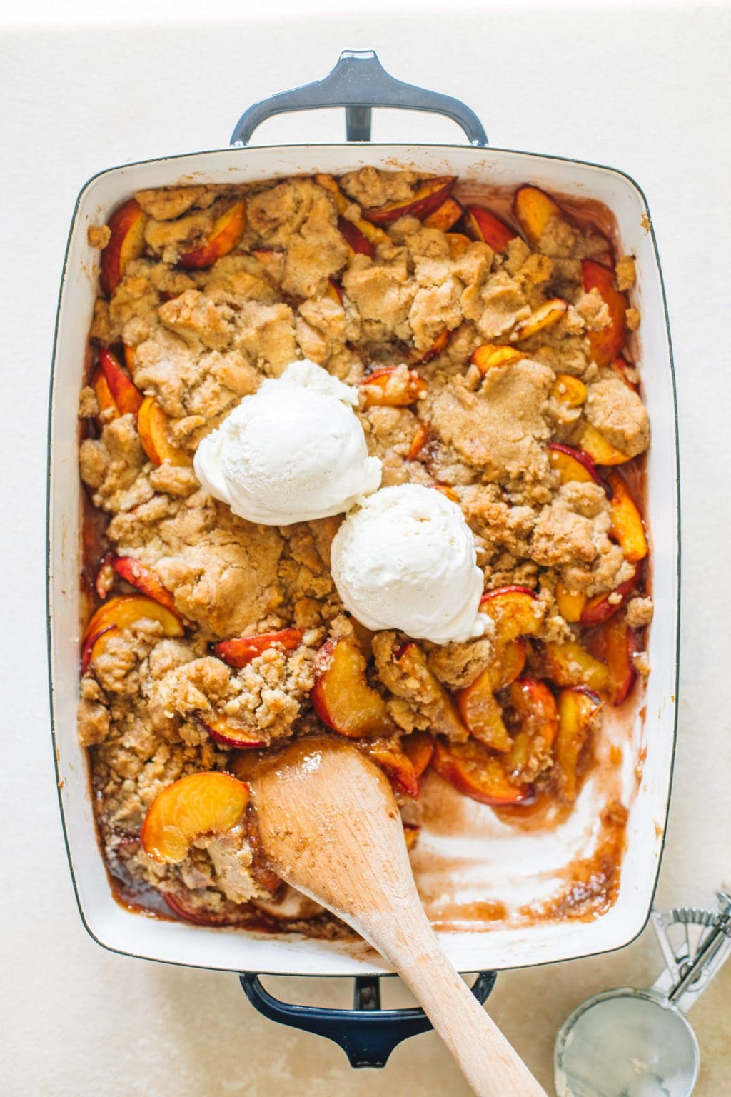 Peach cobbler in a navy blue casserole dish that has been baked and topped with vanilla ice cream.