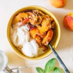 Peach cobbler served in a small yellow bowl with two scoops of vanilla ice cream.