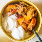 Peach cobbler served in a small yellow bowl with two scoops of vanilla ice cream.