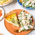 A slice of quiche on a brown plate with a side of everything bagel salad.