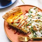 A slice of quiche drizzled with hot sauce, served on a brown plate with a side salad.