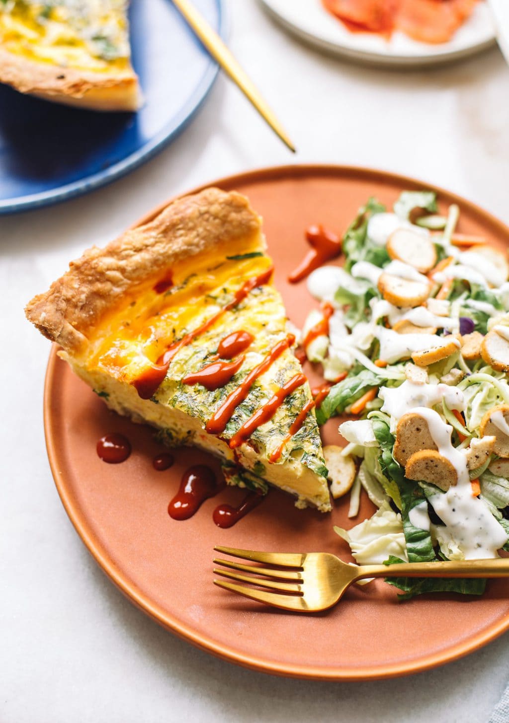 A slice of quiche drizzled with hot sauce, served on a brown plate with a side salad.