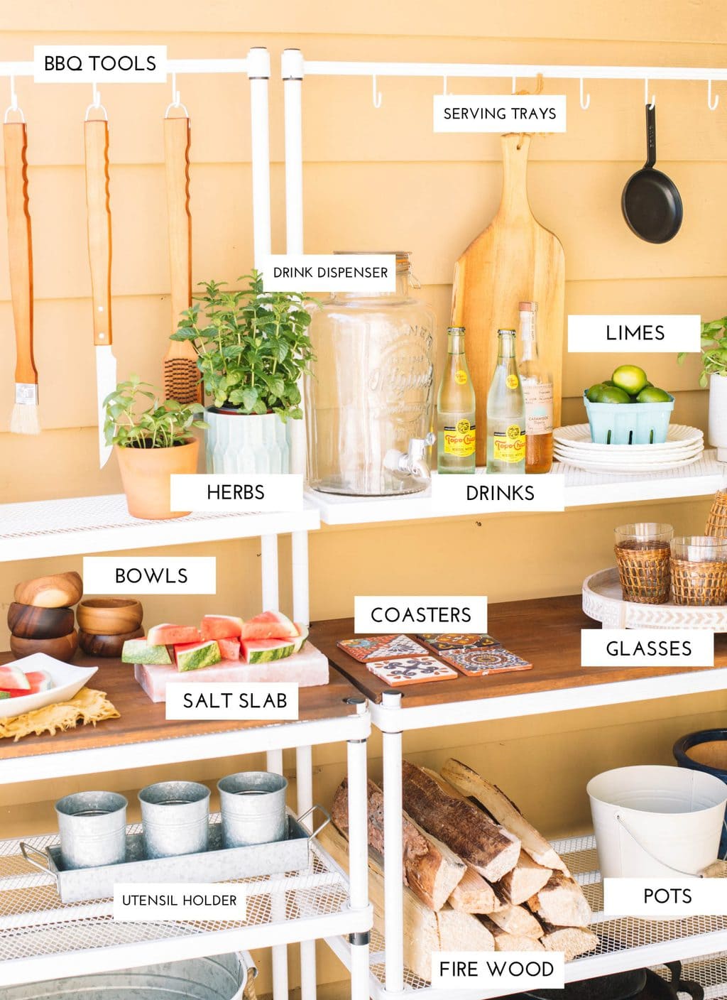 An outdoor buffet set up with a drink dispenser, herbs, glassware, plates, and a utensil holder.