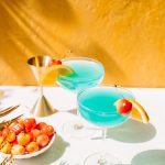 two blue curacao drinks in coupe glasses with orange slice and bowl of cherries