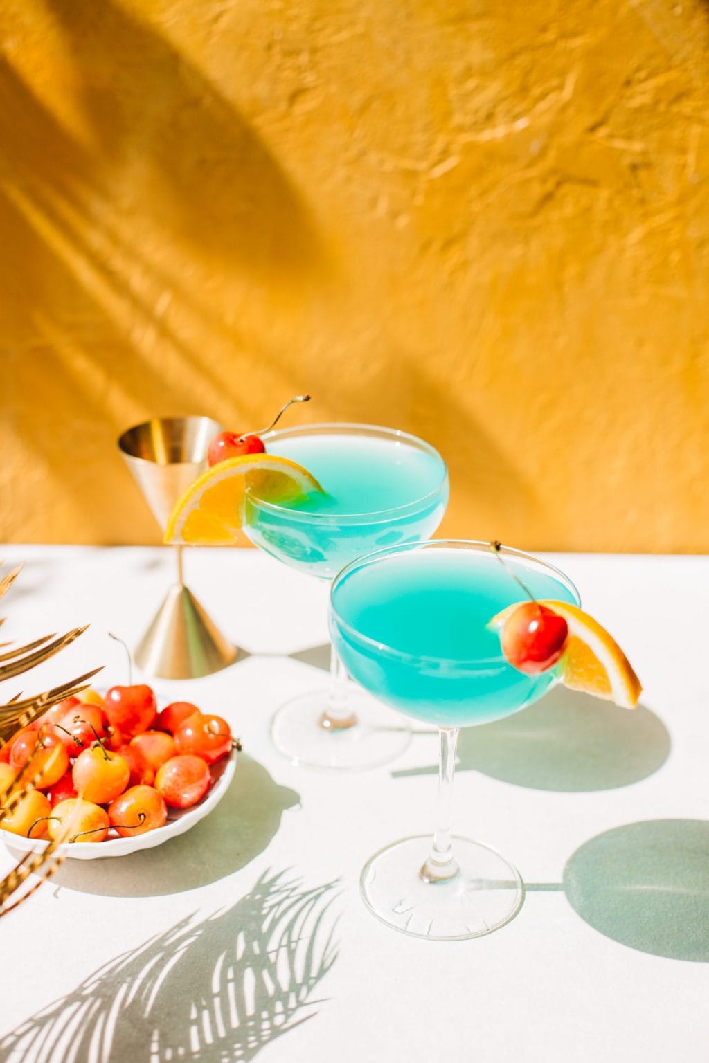 two blue curacao drinks in coupe glasses with orange slice and bowl of cherries