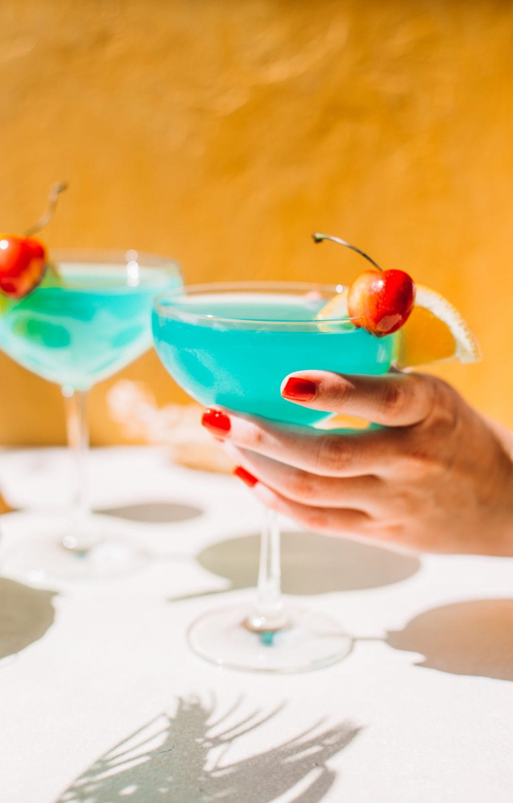 hand holding blue lagoon cocktail in coupe glass with blue caracao drink in background