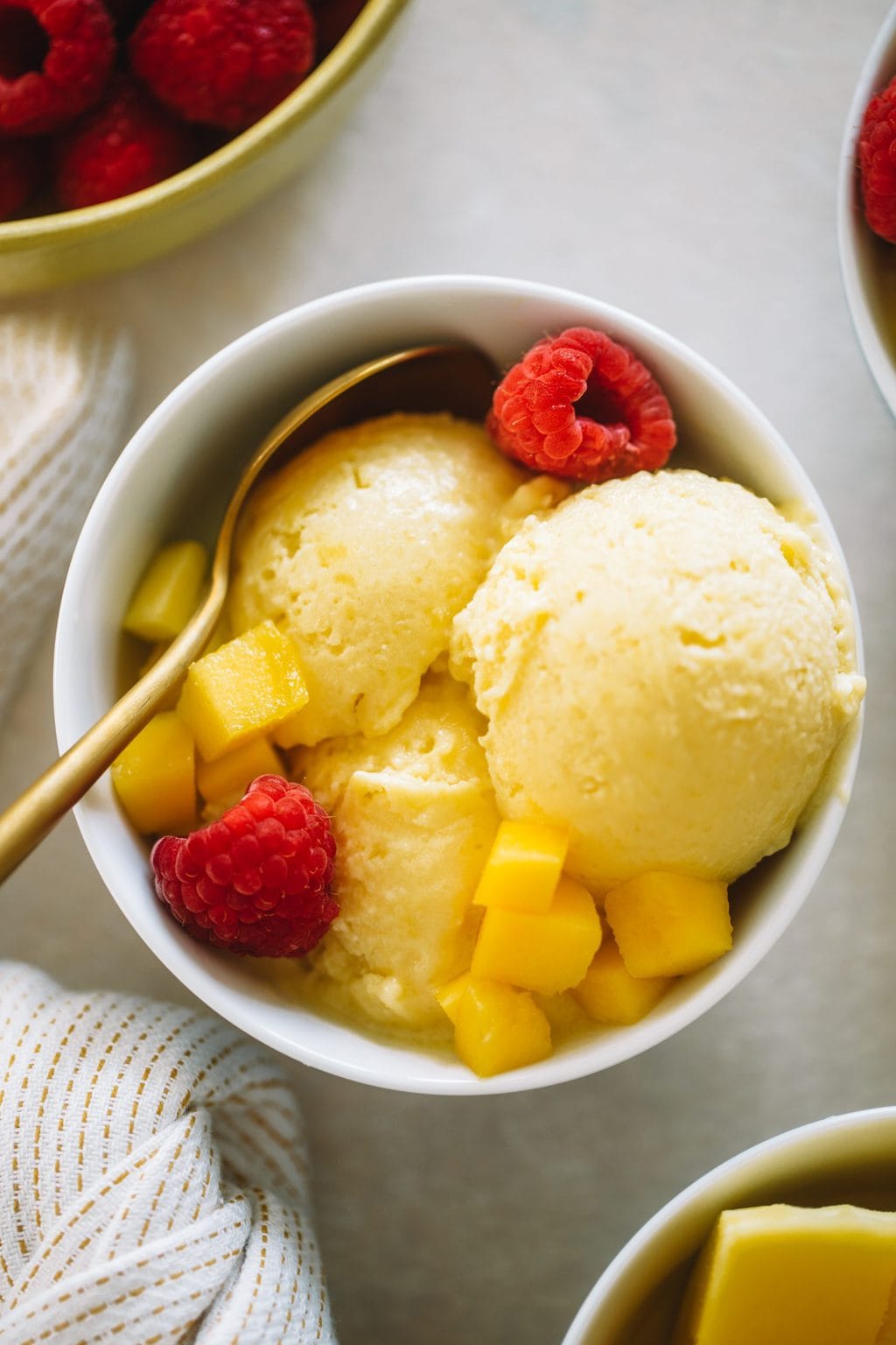 Mango ice cream served in small white bowls topped with fresh raspberries and diced mango.