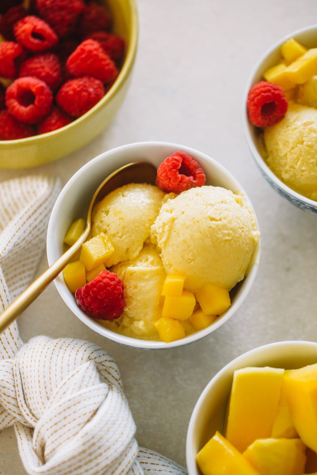 Mango ice cream served in small white bowls topped with fresh raspberries and diced mango.