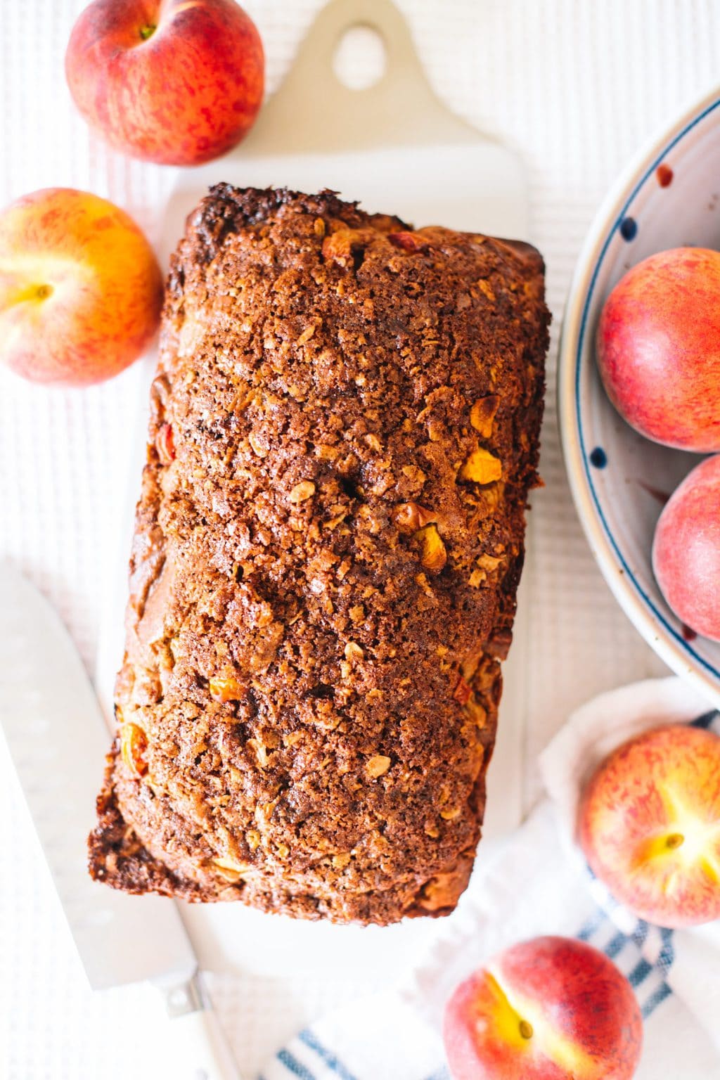 A loaf of peach bread with cinnamon crunch topping and peaches laying next to it. 