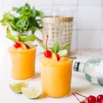 two peach frozen cocktails with maraschino cherry and lime garnish with mint in background