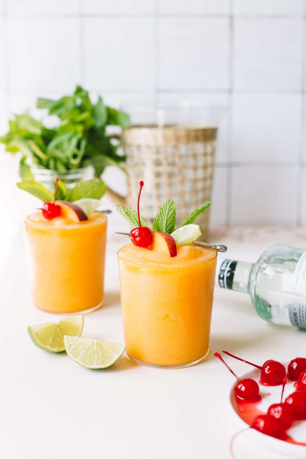 two peach frozen cocktails with maraschino cherry and lime garnish with mint in background