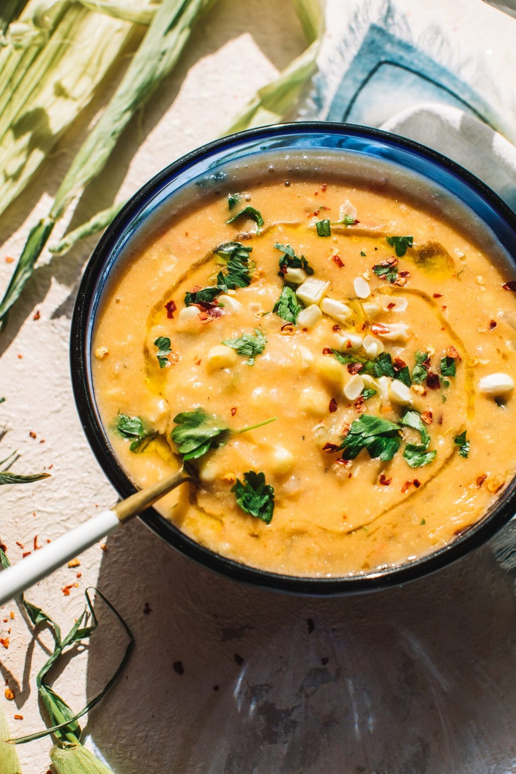 easy corn soup recipe in blue bowl garnished with fresh corn, chives and parsley with large gold spoon