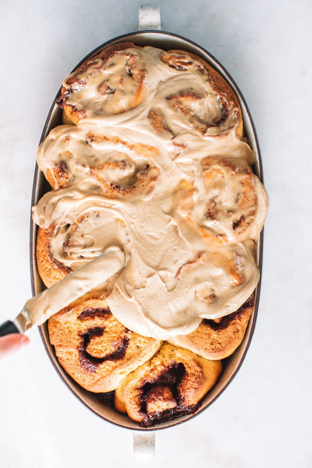 On hand holding a knife and spreading icing over cinnamon rolls in a dish.