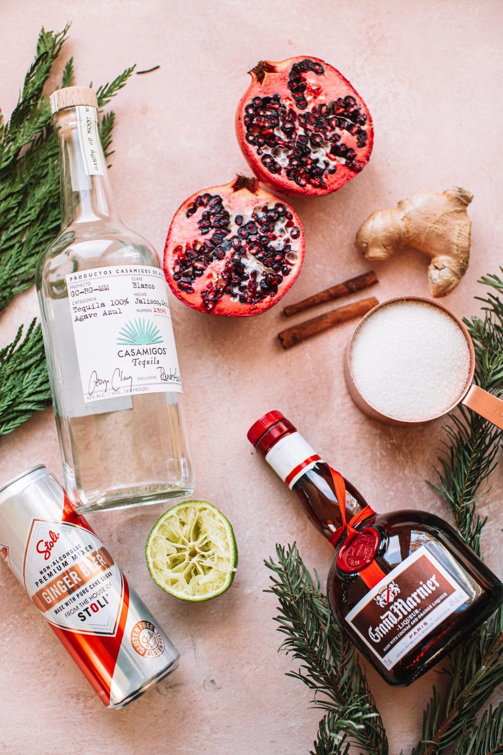 Ingredients for christmas margarita, including a can of ginger beer, a half lime, a glass bottle of tequila, sliced pomegranates, and Grand Marnier liqueur 