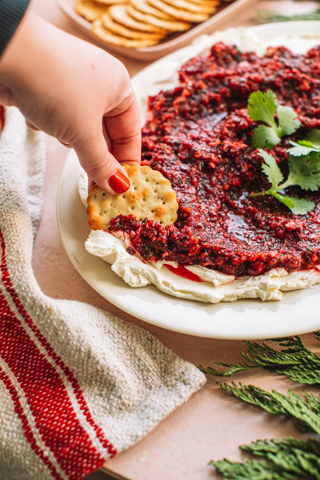 Hand dipping cracker in red jalapeno cranberry dip with cream cheese spread on white plate next to dish of crackers