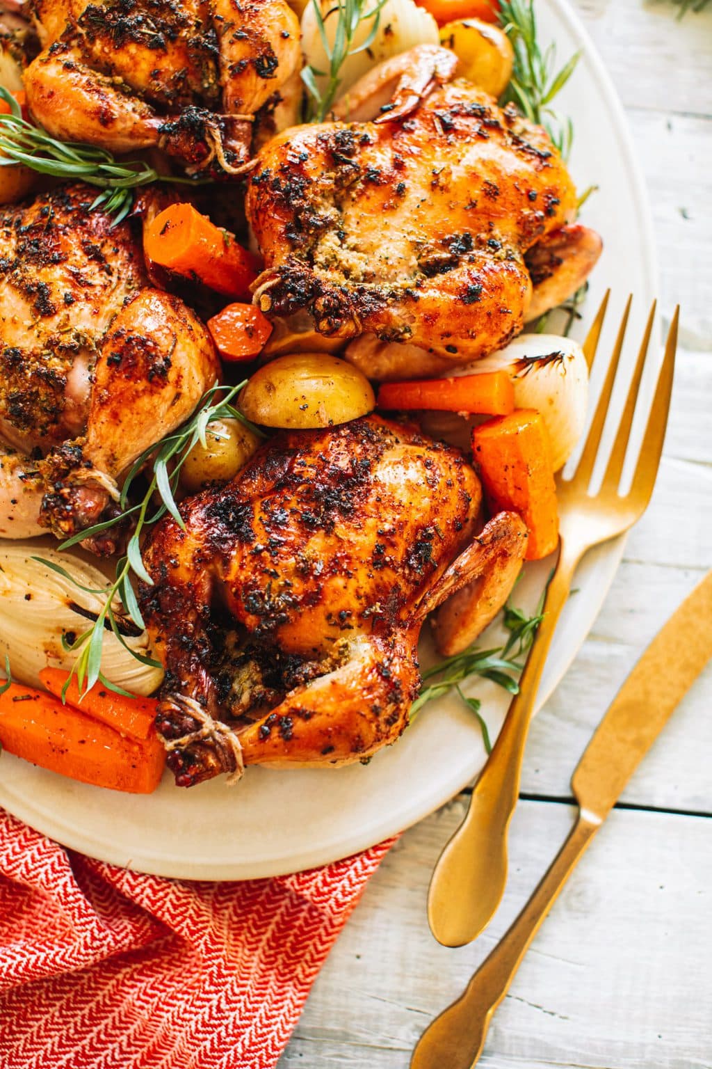 Four oven baked cornish hens arranged on a white plate with carrots, onions and potatoes