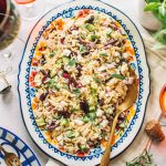 Top shot of greek pasta salad with gold spoon on large oval serving dish.