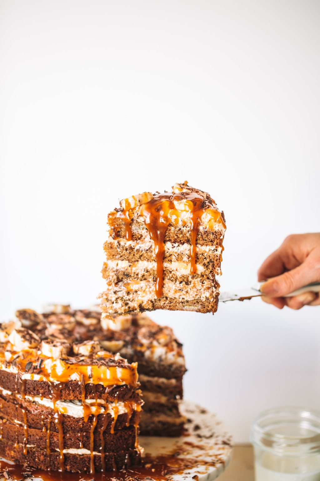 Slice of banana caramel cake being taken from whole cake off of cake stand.