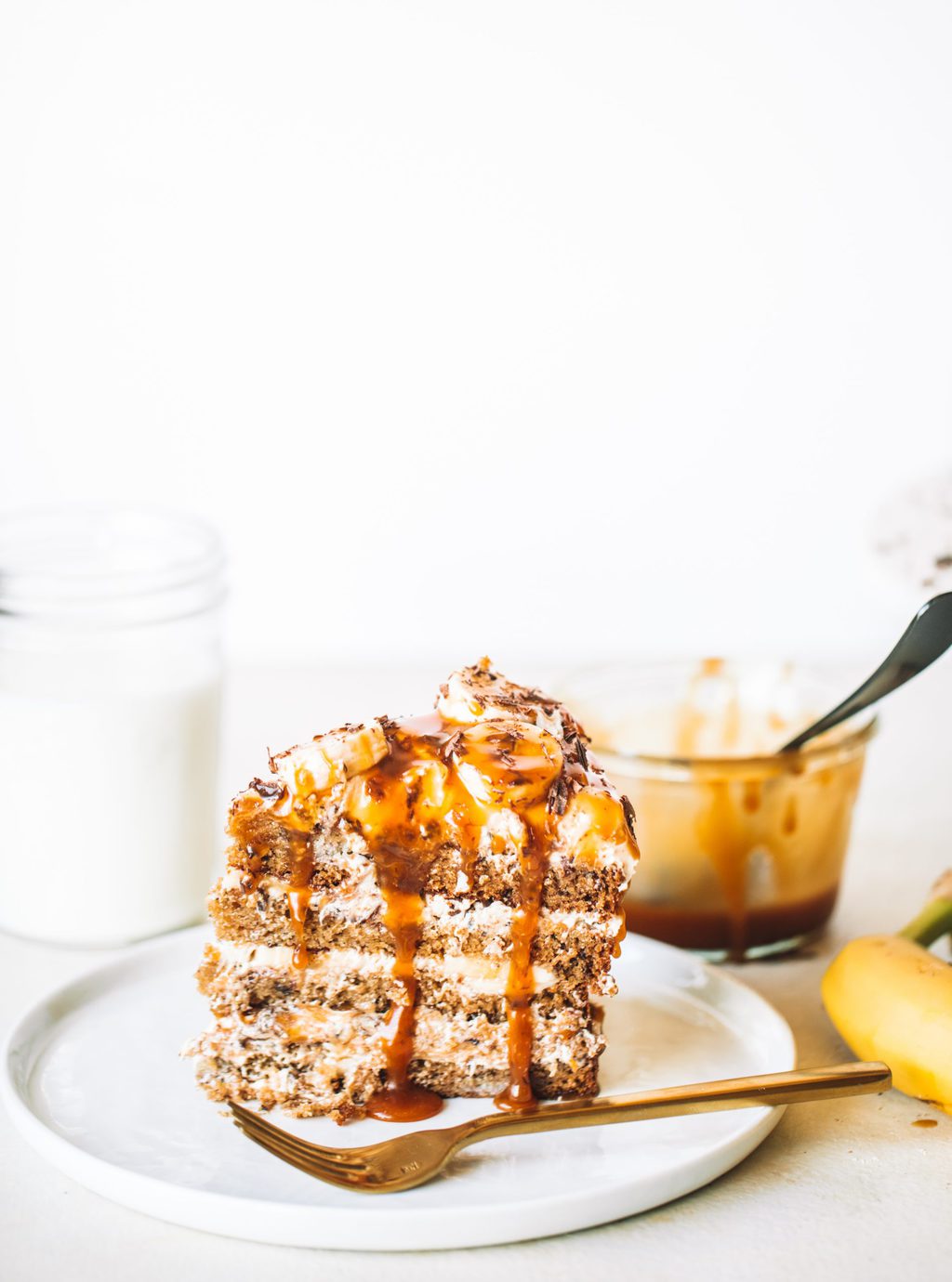 Slice of banana and caramel cake on white plate next to gold fork.