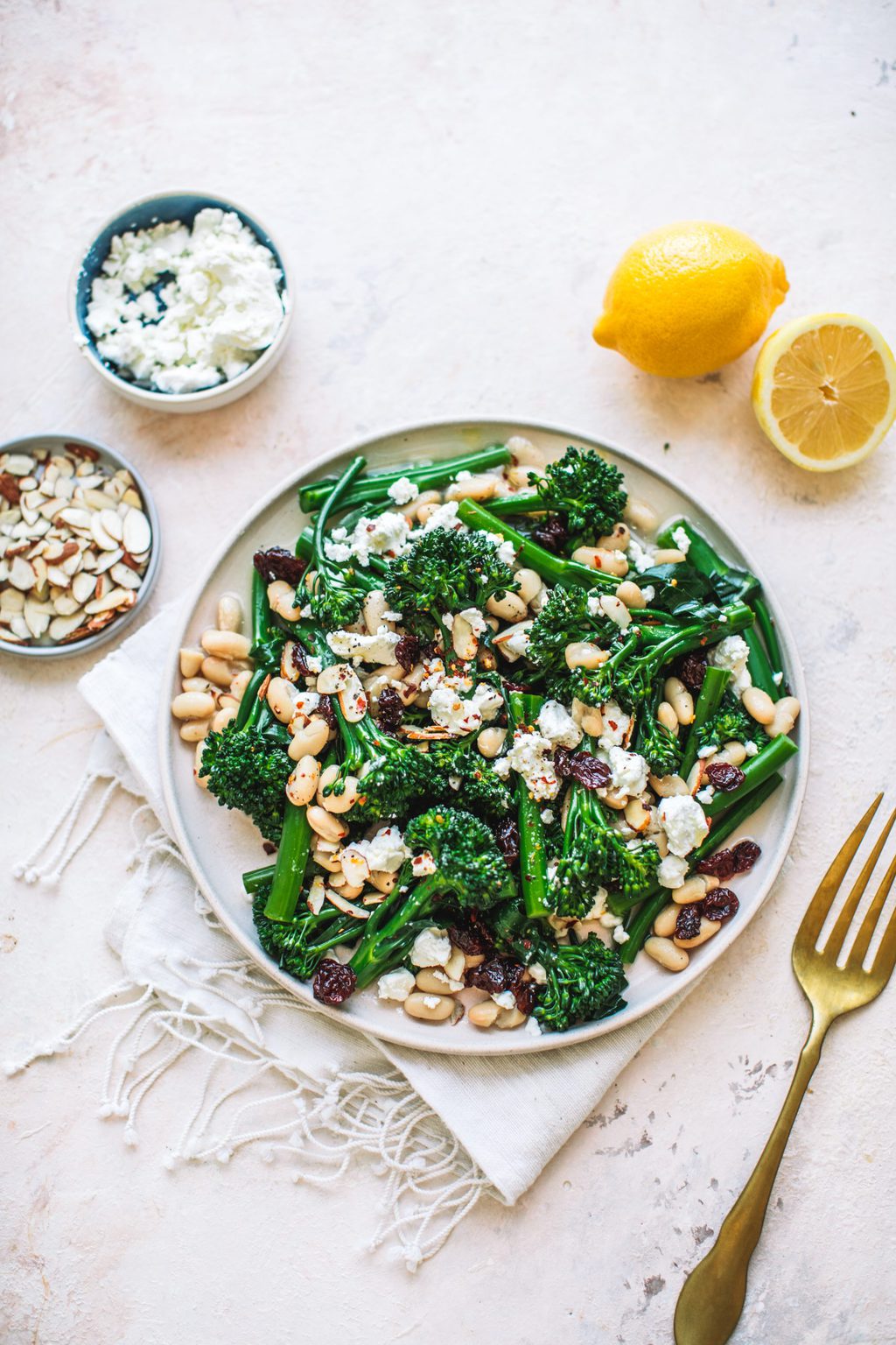 Top shot of broccoli with dried cranberries and white beans on plate next to gold fork and sliced lemon.