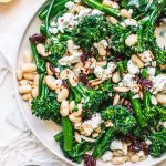 Top shot of broccolini salad with crumbled cheese, toasted almonds and white peans on round white plate.