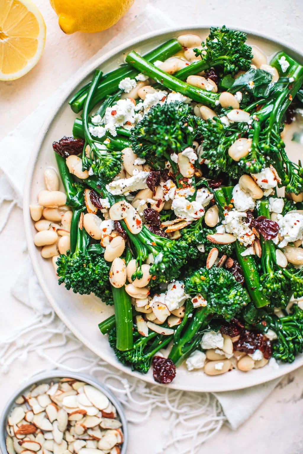 Top shot of broccolini salad with crumbled cheese, toasted almonds and white peans on round white plate.