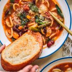 Dipping toast into tomato, vegetable and pasta soup in white bowl.