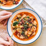 Top shot of two bowls of minestrone soup with gold spoons next to slice of toast and tea towels.