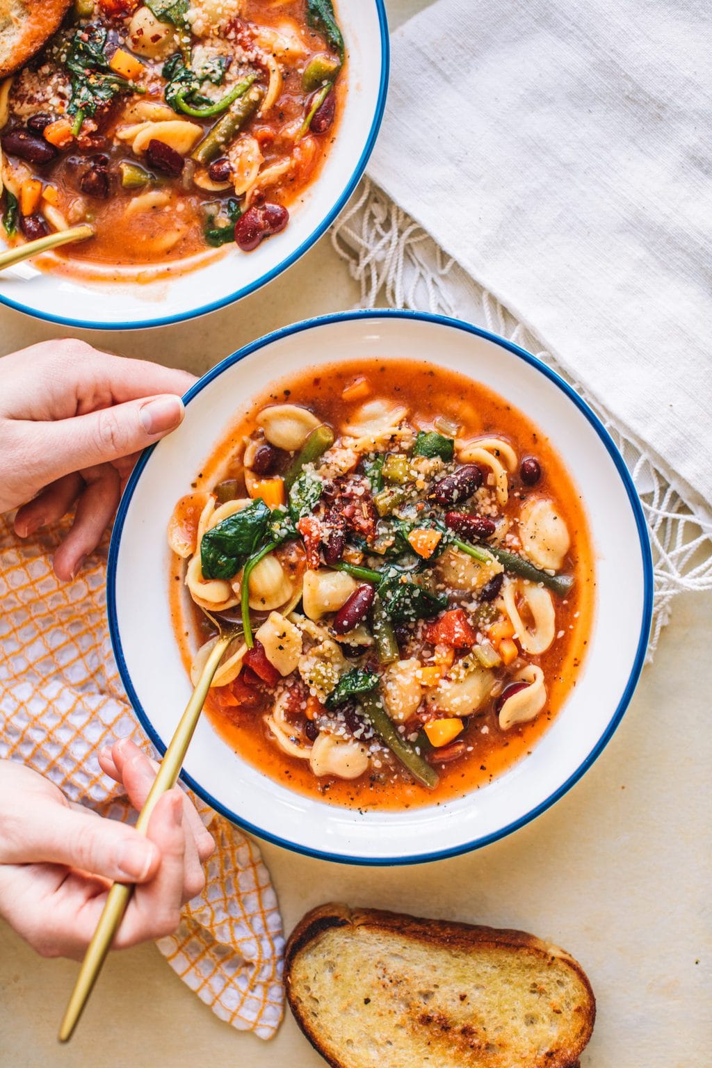 Top shot of two bowls of minestrone soup with gold spoons next to slice of toast and tea towels.