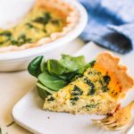 slice of spinach quiche on a plate with fresh spinach and fork