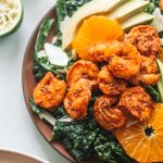 Blackened shrimp on bed of lettuce with avocados and orange slices on red plate.