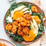 Blackened shrimp on bed of lettuce with avocados and orange slices on red plate.