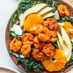 Blackened shrimp on bed of lettuce with avocados and orange slices on red plate.