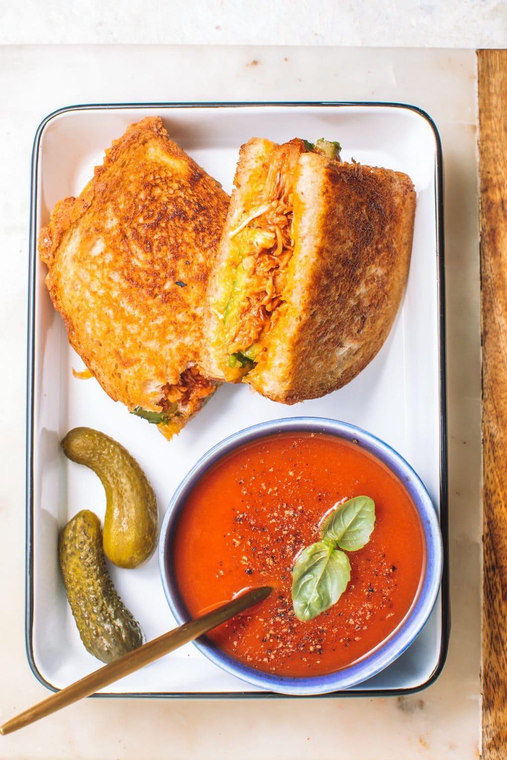 Sandwich served on an enamel platter with a small bowl of tomato soup and two pickles.