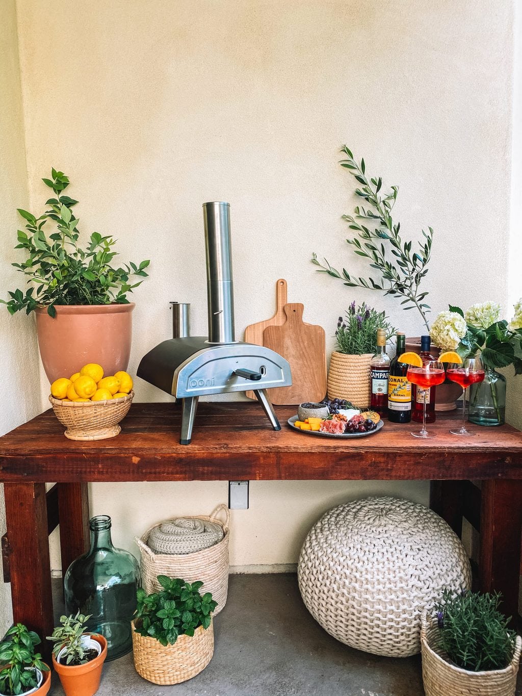 Pizza oven table with a wood fired pizza oven, plants, cutting boards, aperol spritz, and a cheese board.