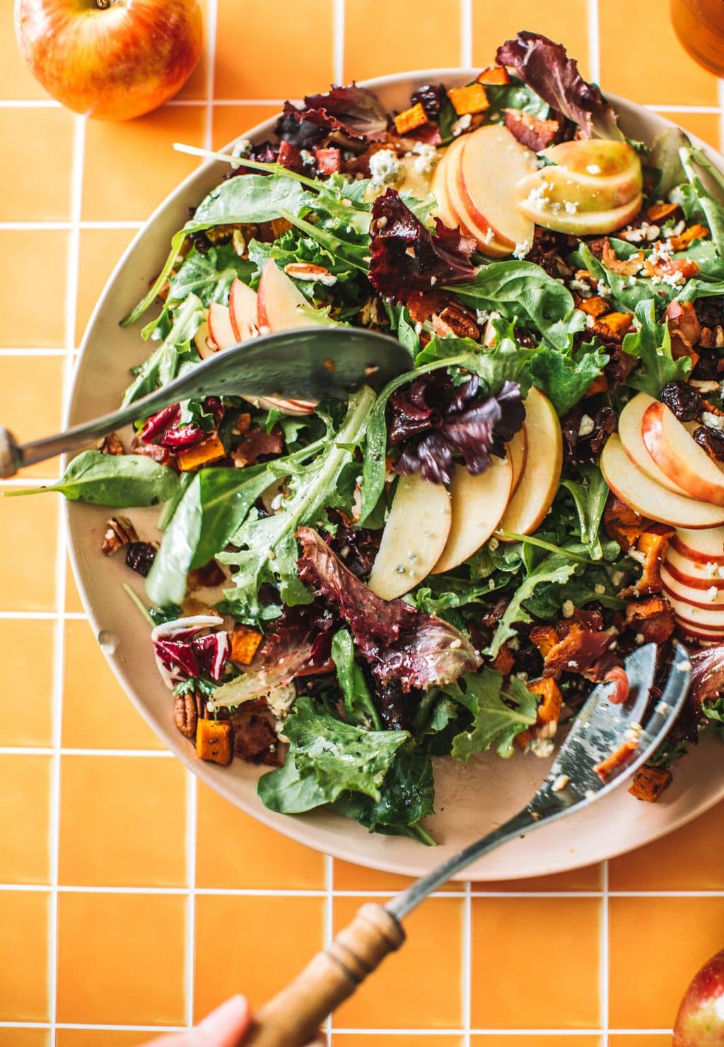 two spoons tossing salad greens, apples, pecans, blue cheese and cranberries in bowl